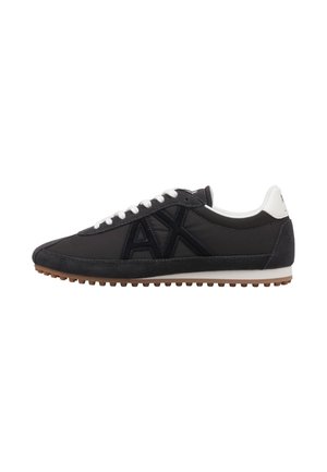 Zapatilla deportiva negra de corte bajo con cordones blancos, gran logo negro "AX" en el lateral, parche blanco en el talón y suela marrón con relieves.