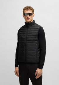 Schwarze gepolsterte Weste mit hohem Kragen, Reißverschluss und strukturierter Oberfläche. Trägt man über einem enganliegenden schwarzen Hemd, kombiniert mit reflektierenden Sonnenbrillen.