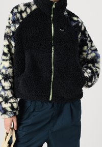 Fleecová bunda s vysokým límcem; černé tělo s vzorovanými, texturovanými rukávy v krémové, modré a zelené barvě; přední zip a boční kapsy.