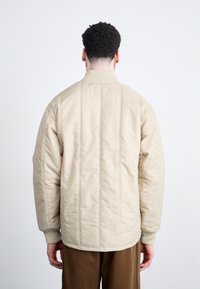 Beige gesteppte Jacke mit gerippten Bündchen und Kragen, mit einer einheitlichen Textur und minimalen Nähten, getragen über braunen Hosen, von hinten betrachtet.