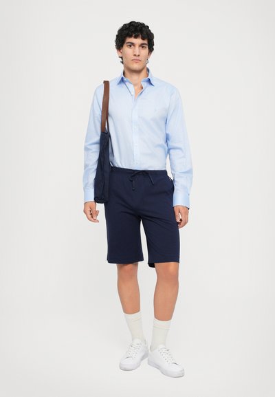 Ljusblå button-up skjorta, marinblå shorts med elastisk midja, vita sneakers och krämfärgade strumpor. Modell som håller i en mörk axelväska.