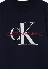 Granatowa tkanina z dużymi białymi literami "cK" oraz czerwonym napisem "CALVIN KLEIN JEANS" na środku odzieży.