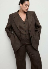 Abito in lana marrone con giacca sartoriale dotata di ampi rever e gilet con chiusura a bottoni. I pantaloni sono coordinati per colore e tessuto.