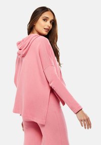 Pink hooded sweatshirt with a relaxed fit and long sleeves, featuring a soft texture and side slits for comfort. Paired with matching pants.