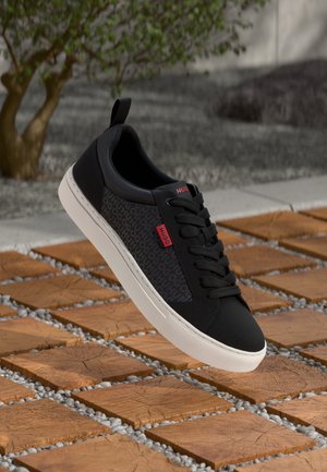 Black sneaker with textured side panel, white sole, and red logo accent. Features round laces and a pull tab on the heel.