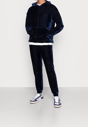 Mężczyzna ubrany w granatową welurową bluzę z kapturem i dopasowane spodnie dresowe, w zestawie z białymi i granatowymi sneakersami, stoi na tle jednolitego tła.