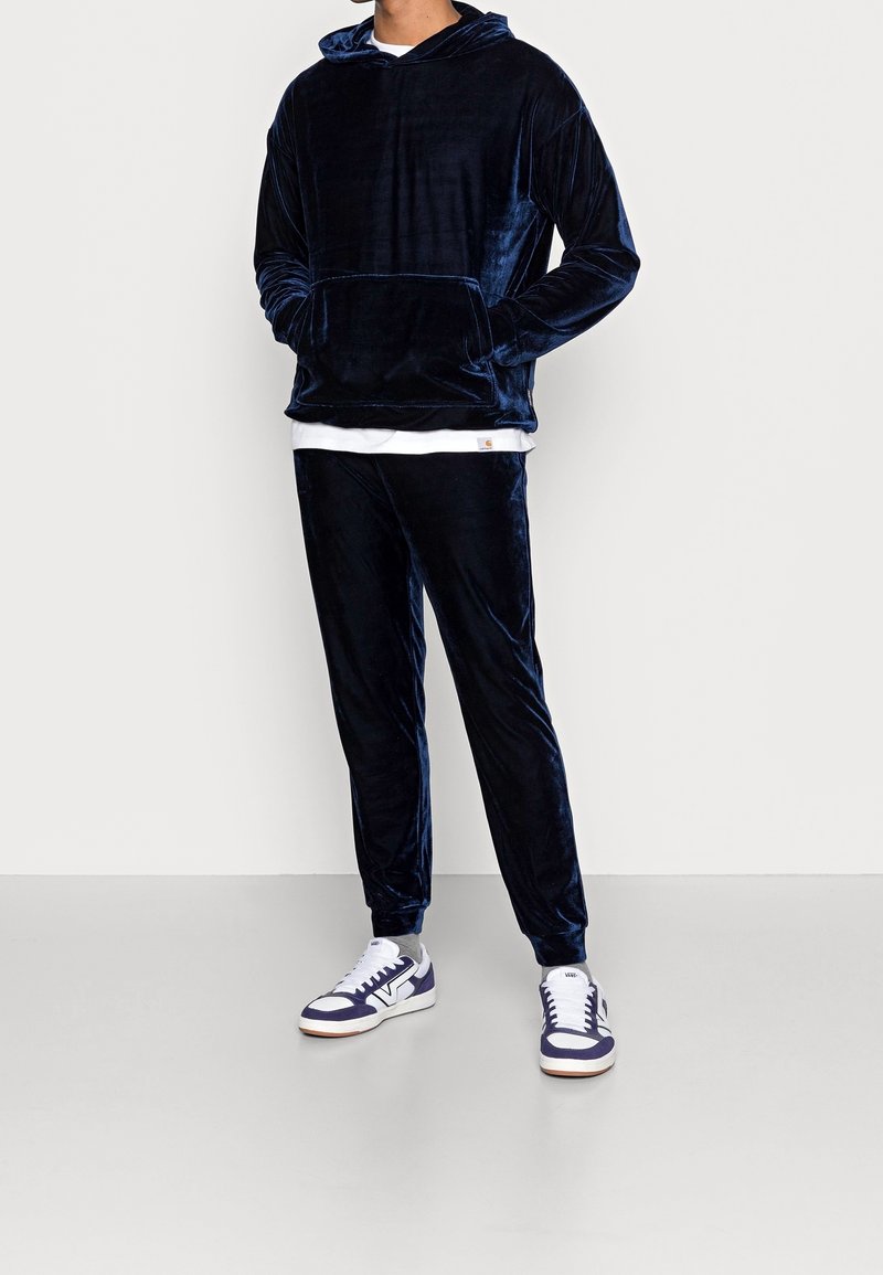Homme portant un sweat à capuche en velours bleu marine avec un pantalon de jogging assorti, accompagné de baskets blanches et bleues marine, debout devant un fond uni.