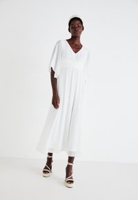 Λευκό φόρεμα μέχρι το γόνατο με φτερούγες και εφαρμοστή μέση. Απαλή ύφανση, V-λαιμόκοψη, συνδυασμένο με μπεζ ψηλοτάκουνα σανδάλια με λουράκια.