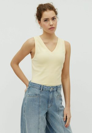 Mladá žena s kučeravými vlasmi v krémovom bezrukávovom topu s výstrihom do V a voľných modrých rifle s vysokým pásom, stojaca s jednou rukou na páse.