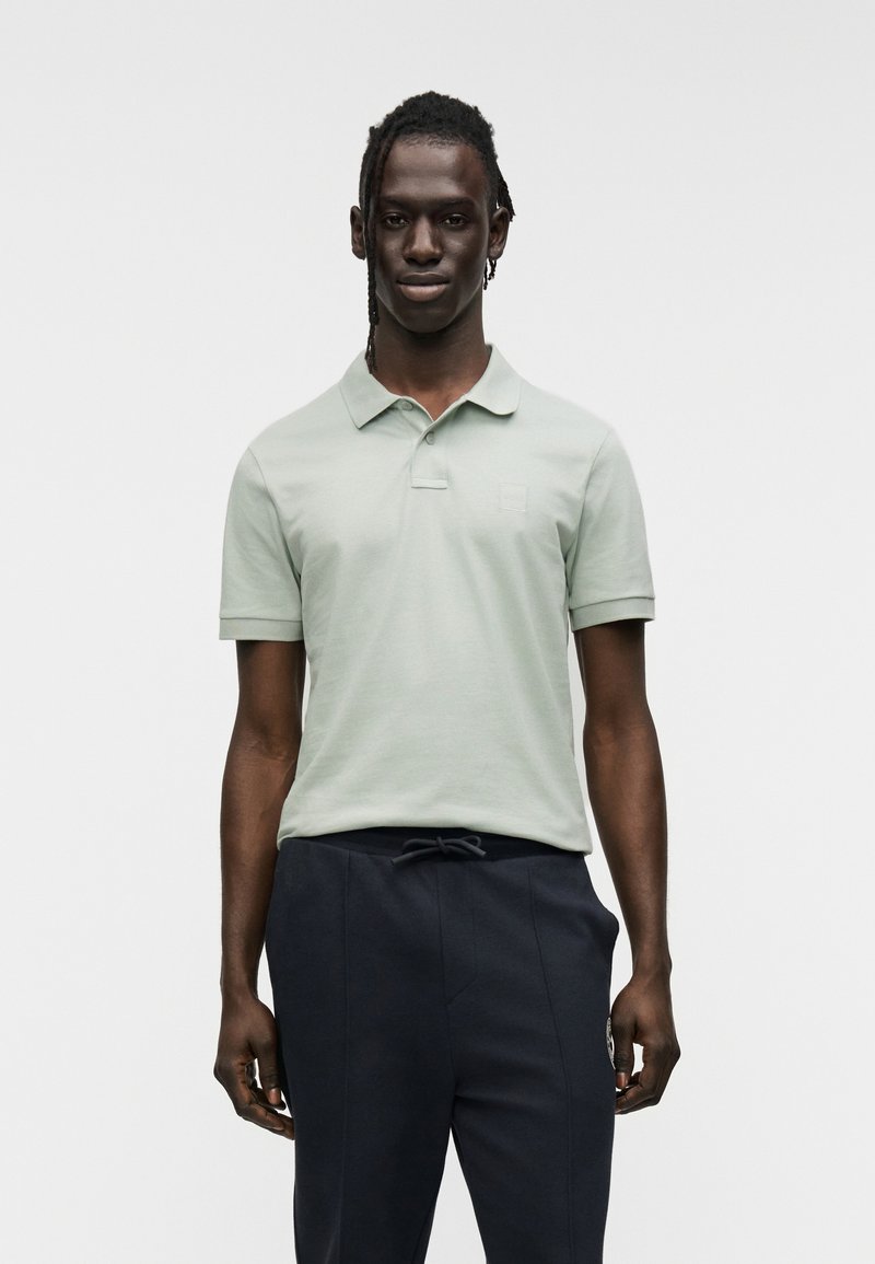 Ανοιχτό πράσινο πόλο με γιακά και κοντά μανίκια, με διακριτικό λογότυπο στο στήθος. Φορεμένο με σκούρο παντελόνι jogger.