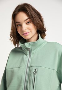 Giacca zip-up verde chiaro con colletto alto, dotata di due tasche frontali e dettagli sul cursore a forma di cuore. Tessuto morbido e testurizzato.