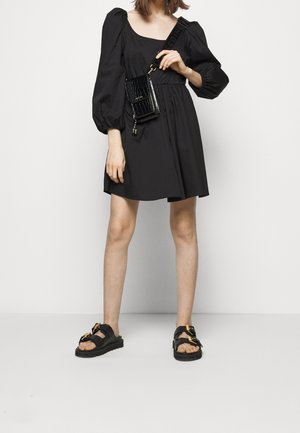 Zwarte jurk met pofmouwen en vierkante halslijn, zachte textuur en een taillering, gecombineerd met zwarte sandalen met gouden accenten.