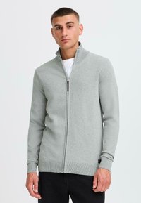 Sudadera de cremallera en gris claro, hecha de un tejido texturizado. Presenta un cuello alto y puños y dobladillo acanalados, llevada sobre una camiseta blanca.