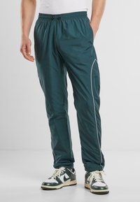 Pantalones deportivos en color azul verdoso con una franja blanca en el costado, cintura elástica y una textura suave, combinados con zapatillas verdes y blancas.