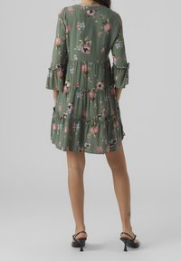 Robe fleurie en vert olive avec des volants superposés. Manches trois-quarts avec des poignets à franges. Imprimée de fleurs roses et bleues.