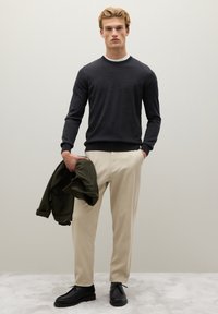 Maglione a maglia grigio scuro, pantaloni crema, scarpe nere. Tiene una giacca verde. Design minimalista con collo rotondo e polsini aderenti. Texture liscia.