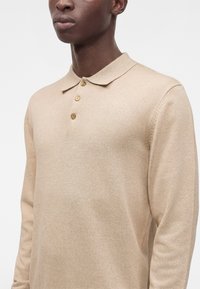 Homme portant un pull polo beige à manches longues avec trois boutons, montré de la poitrine au cou sur un fond blanc uni.
