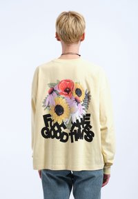 Langärmliges gelbes T-Shirt mit einem floralen Motiv, das Sonnenblumen und Mohnblumen zeigt, kombiniert mit kräftigem Text "FÜR DIE GUTEN ZEITEN."