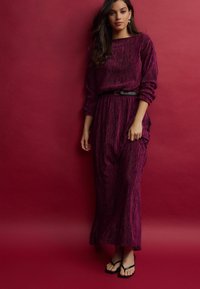 Robe longue de couleur prune avec un design texturé et plissé, dotée de manches longues et d'une taille cintrée avec une ceinture noire. Assortie de tongs noires.