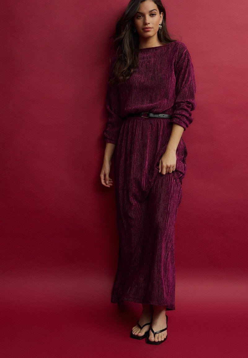 Robe longue de couleur prune avec un design texturé et plissé, dotée de manches longues et d'une taille cintrée avec une ceinture noire. Assortie de tongs noires.