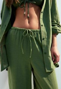 Groene linnen top en bijpassende broek. De top heeft een trekkoord en zilveren accenten; de broek heeft een elastische tailleband met een strik. Textuurstof.