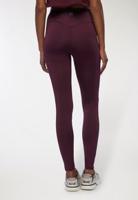Leggings bordeaux realizzati in tessuto elasticizzato, con una vita alta e una texture liscia. Abbinati a scarpe sportive grigie.