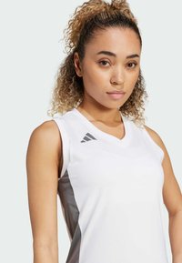 Top deportivo sin mangas de color blanco con escote en V, que presenta paneles laterales grises y un logo de tres franjas grises en el hombro izquierdo.