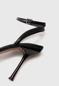 Sanda con tacco alto nera in finitura verniciata lucida, con una sottile cinghia alla caviglia e design a punta. Caratterizzata da un tacco stiletto snello.