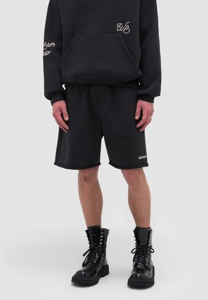 Person wearing black hoodie with front pocket, black shorts with small text, and black lace-up combat boots against gray background.