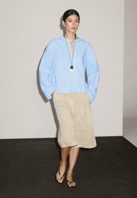 Une femme se tient avec confiance dans un chemisier bleu clair et une jupe beige, portant un long collier et des sandales noires, sur un fond neutre.