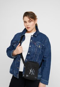 Borsa a tracolla nera dei New York Yankees con logo argento, abbinata a una giacca di jeans blu e una maglietta bianca. Tessuto morbido, design compatto.