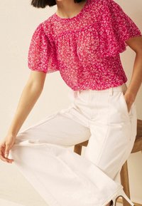 Blouse en dentelle florale rose avec des manches bouffantes, associée à un pantalon large taille haute blanc. L'ensemble présente des détails délicats et des contrastes de textures.