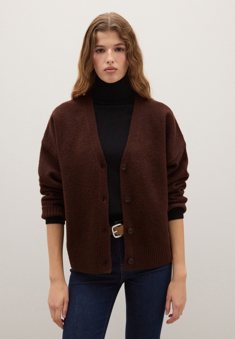 Brauner, übergroßer Cardigan mit weicher Textur, gerippten Bündchen und fünf Knöpfen. Getragen über einem schwarzen Rollkragenpullover und kombiniert mit blauen Jeans.