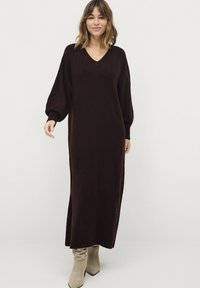 Lang, donkerbruin gebreid jurk met een V-hals, ballonmouwen en geribbelde zoom. Gecombineerd met lichtbeige haklaarzen.