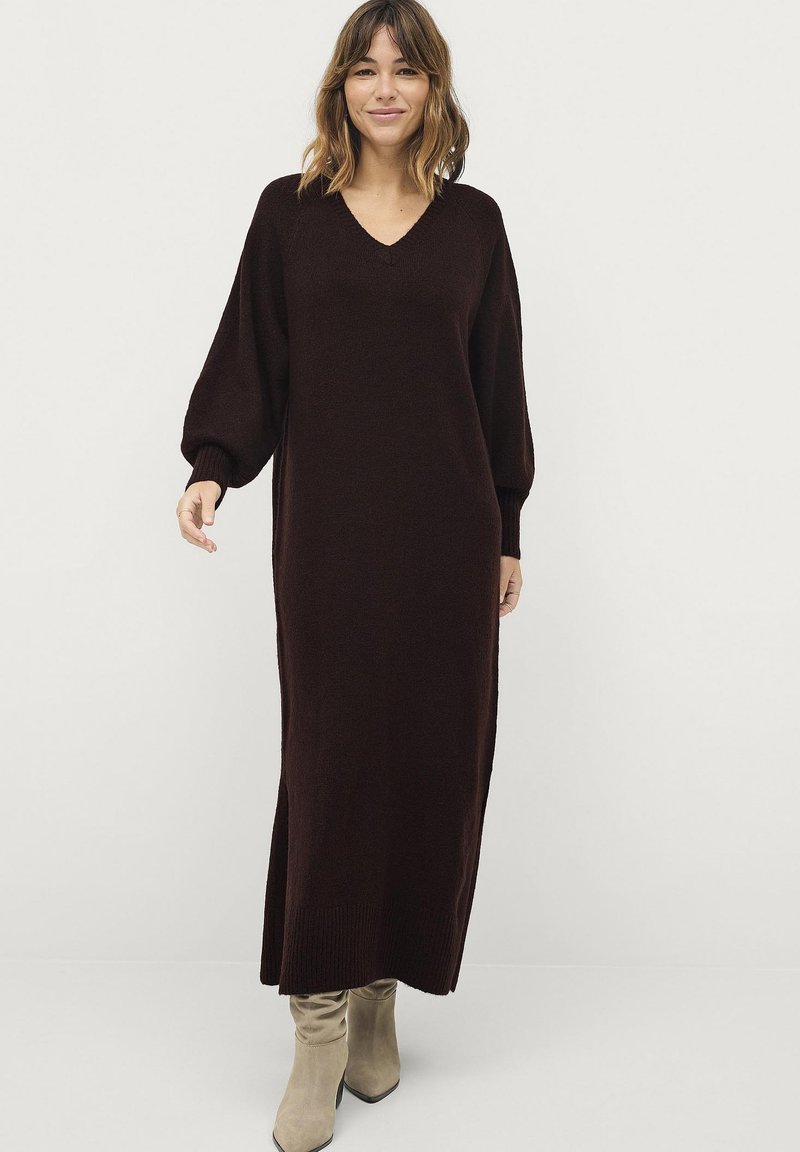 Lang, donkerbruin gebreid jurk met een V-hals, ballonmouwen en geribbelde zoom. Gecombineerd met lichtbeige haklaarzen.