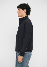 Veste marine en tissu lisse, avec un col montant, deux poches latérales et des poignets côtelés. Associée à un jean bleu clair.