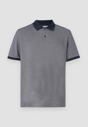 Homme debout portant un polo gris col noir, un pantalon bleu marine et des sandales noires à enfiler, sur un fond clair uni.