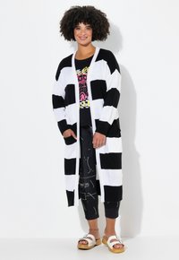 Cárdigan largo a rayas blanco y negro con abertura frontal, puños acanalados y dos bolsillos, superpuesto sobre una camiseta gráfica negra y pantalones desgastados.
