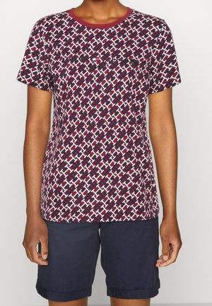 Personne portant une chemise à manches courtes bordeaux et blanche à motif avec un short bleu marine sur un fond clair uni.
