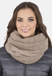 Écharpe infinity tricotée en beige doux, présentant une texture épaisse et un design en boucle, portée sur une veste grise matelassée.