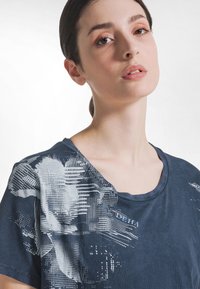 Młoda kobieta o jasnej cerze i ciemnych włosach ma na sobie granatową koszulę z białymi abstrakcyjnymi wzorami na ramieniu i klatce piersiowej.