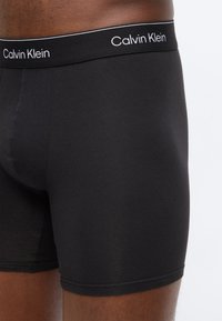 Nærbilde av en person som har på seg svarte Calvin Klein boxer shorts med en merkevaremerket linning mot en ensfarget bakgrunn.