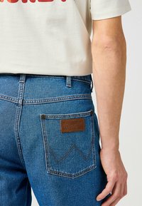 Blauwe denim jeans met een bruine leren Wrangler-patch op de achterzak, met contrasterende stiksels en een klassiek fit ontwerp.