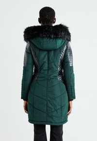 Pildīta garā mētelis tumši zaļā krāsā ar noņemamu melnu mākslīgās kažokādas kapuci, ar diognālām kviltējuma rakstēm un kontrastējošām tekstūrām piedurknēm.