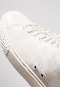 Gros plan d’une basket en cuir blanc montrant une surface texturée, des lacets blancs, des œillets métalliques et une semelle en caoutchouc épaisse avec une semelle à motif.