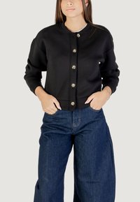 Felpa nera con scollatura rotonda, chiusura frontale con bottoni in metallo argentato e polsini a coste. Abbinata a pantaloni in denim larghi.