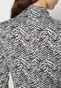 Nahaufnahme einer Person, die ein schwarz-weiß gestreiftes Turtleneck-Oberteil mit einem kleinen goldenen, insektförmigen Anstecker am hinteren Kragen trägt.