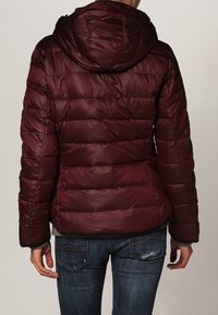 Veste matelassée bordeaux avec capuche, présentant un design matelassé, des poignets élastiques et un ourlet au niveau des hanches. Portée avec un jean bleu.