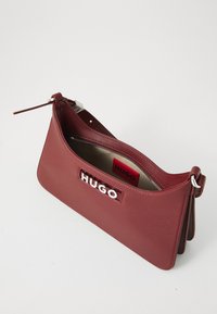 Pravokutna tašna od burgundske kože s srebrnim HUGO logotipom, gornjim zatvaračem i podesivim remenom na bijeloj pozadini.
