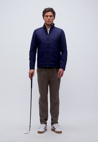 Νεαρός άνδρας που στέκεται, κρατώντας ένα μπαστούνι γκολφ στο δεξί του χέρι, φορώντας ένα ναυτικό μπουφάν, μπεζ παντελόνι και λευκά αθλητικά παπούτσια.